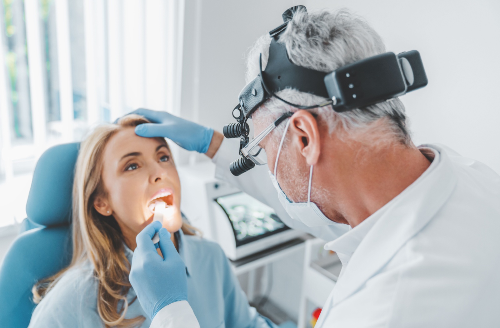 A dentist holds a patient’s forehead while opening their mouth with a popsicle stick to examine their mouth